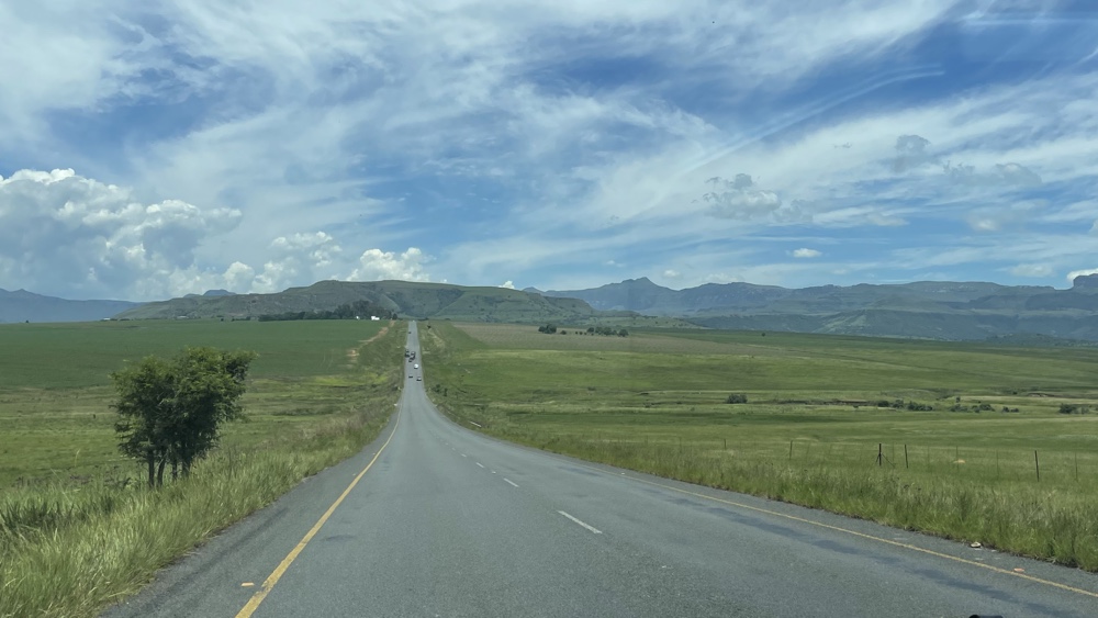 A straight road with fileds on either side and mountains in the distance.